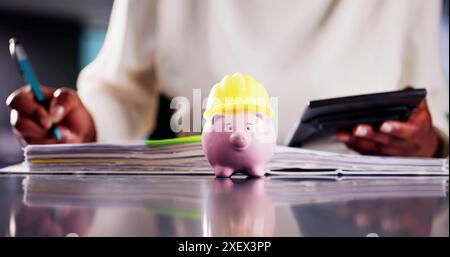 Young African-American woman in hard hat crunches numbers in office, saving money with piggy bank and managing bank invoices. Stock Photo