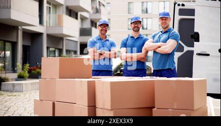 Young and happy male movers in protective suits standing in front of van, delivering boxes for a moving service company. Stock Photo