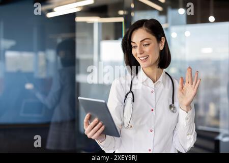 Young female doctor using tablet for video call in modern office. Professional in white coat with stethoscope having virtual consultation, connecting with patients online, waving at screen Stock Photo