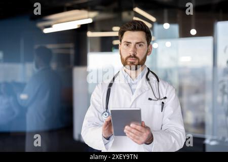 Doctor in white coat with stethoscope using digital tablet in hospital. Medical professional focused on patient data and healthcare technology. Office background with blurred colleagues. Stock Photo