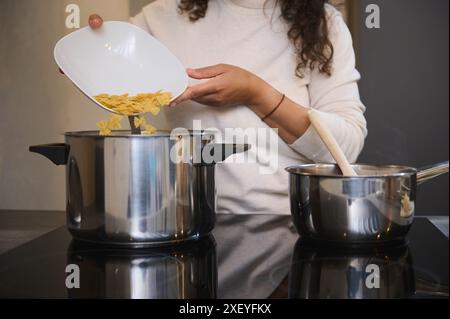 Details on the hands of a woman chef pouring raw farfalle macaroni into a boiling water in the stainless steel pan, cooking Italian pasta in the home Stock Photo