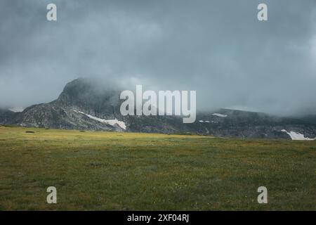 Fog covering the peak with snow leftovers during summer. Cold rainy weather. Vibrant green meadow. Stock Photo