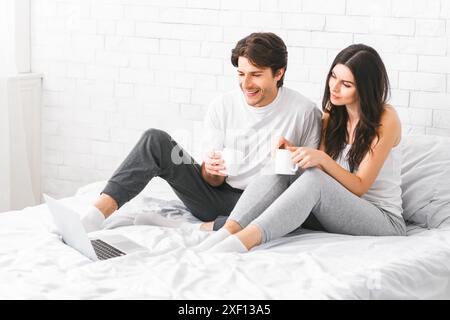 Couple Sitting On Bed Drinking Coffee And Using Laptop Stock Photo