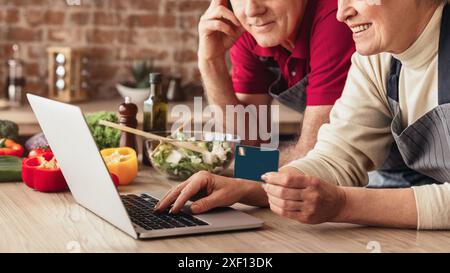 Senior Couple Using Laptop and Credit Card in Kitchen, Cropped Stock Photo