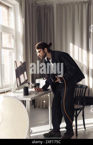 Vertical full length portrait of young professional photographer holding camera and using computer in minimal photo studio with light accents  Stock Photo
