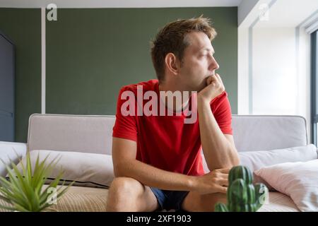 Thoughtful man in a red shirt sits on couch at home and looking in the window. Tired bored male sitting on sofa in modern living room. Stress Stock Photo