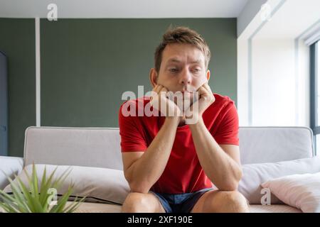 Bored sad man in a red shirt sits on couch at home with his chin resting on his hands. Tired pensive male sitting on sofa in modern living room Stock Photo