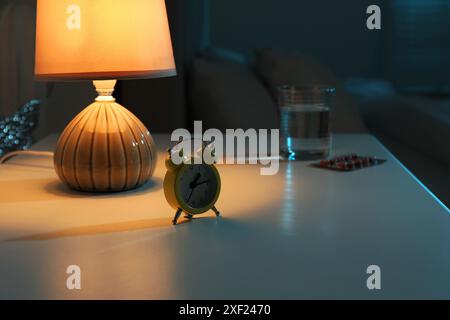 Insomnia treatment. Glass of water, pills and alarm clock on bedside table in bedroom at night, selective focus Stock Photo