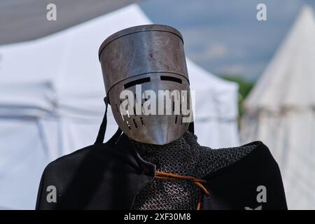 A knight in a full-face metal helmet and chainmail stands in front of medieval tents. The armor is detailed, featuring a slit visor and ventilation ho Stock Photo
