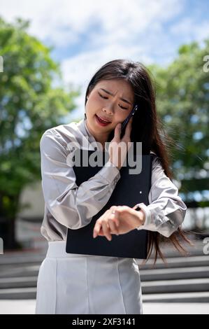 A busy, frustrated Asian businesswoman with a file folder is talking on the phone with her colleague and checking the time on her wristwatch while hur Stock Photo