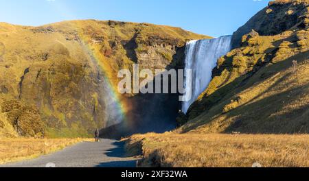 A rainbow is seen in the sky above Skogafoss waterfall. The scene is serene and peaceful, with the water cascading down the rocks and the rainbow addi Stock Photo