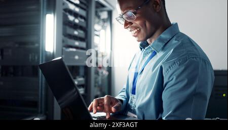 Laptop, server room and black man on database network for maintenance, programming software or typing. Tech, data center and happy engineer on system Stock Photo