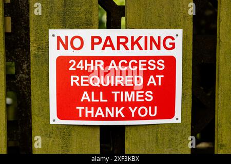 a red and white warning sign for no parking, with 24HR access required at all times fixed to a wooden fence Stock Photo