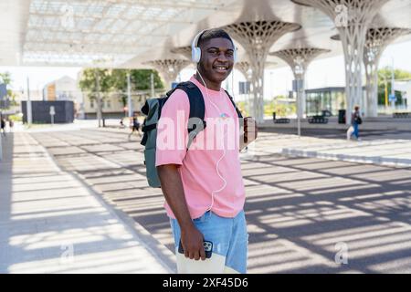 A young african man wearing a pink shirt and jeans is smiling and holding a cell phone. He is wearing headphones and has a backpack on. The scene take Stock Photo