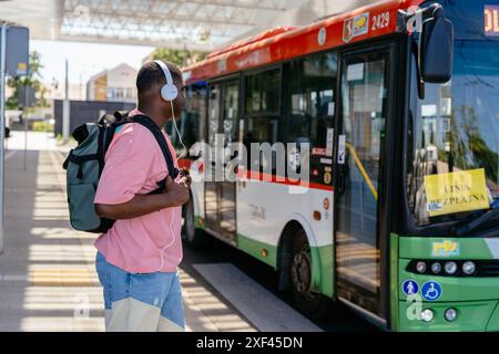 African man wearing headphones and a backpack stands next to a green and white bus. Handsome black teen guy with backpack boarding bus in city. Stock Photo