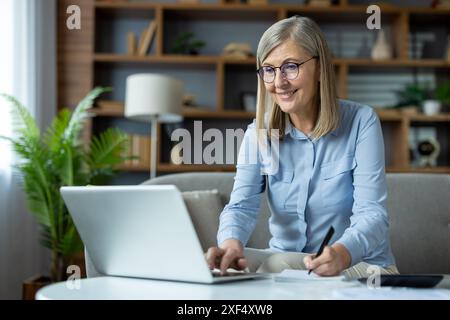 Smiling senior woman working on a laptop at her home office. She's taking notes and appears focused and happy. Perfect for concepts of remote work, home office, senior professionals, and technology Stock Photo