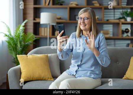 A smiling senior woman with gray hair and glasses is video chatting on a smartphone while sitting on a sofa in a cozy, modern living room with wooden shelves and indoor plants. Stock Photo