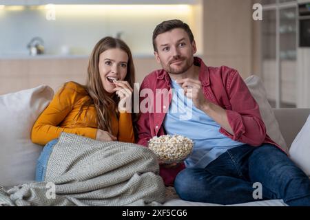 Cheerful married couple, spouses watching funny movie, series relaxing on couch, eating popcorn. Stock Photo