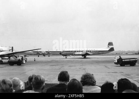 Germany Circa. 1960 – A group of spectators watching aircraft at an airport. In the foreground is the Vickers Viscount 814 turboprop airliner of the German airline, Deutsche Lufthansa; registration, D-ANEF. The aircraft is decorated in its original livery. This aircraft was in service with Lufthansa from March 1959 until 20 January 1972, when it was purchased by Airwork Services Ltd. Eight days later, the aircraft was damaged beyond repair during the delivery flight, after it made a heavy landing at Hurn Airport, Bournemouth, England. An airport worker is driving a MIAG Plattenwagen vehicle. Stock Photo