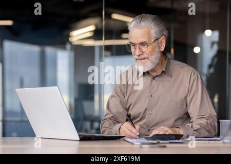 Senior businessman working on a laptop and taking notes in a modern office. The image captures a mature professional in a work environment, showcasing productivity, experience, and dedication Stock Photo
