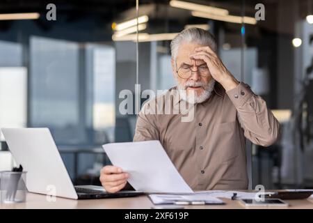 Senior businessman looking stressed while reading documents at his office desk. He is sitting in front of a laptop, holding a paper and appears to be in deep thought or confusion. Stock Photo