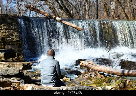 Visitor to the Amis Mill Ruins and Dam, Sits and watches the water spill over the dam.  He is wearing a grey jacket. Stock Photo