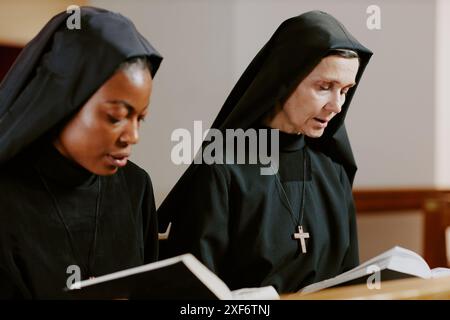 Medium closeup of two ethnically diverse young and mature Catholic nuns praying while sitting on pew Stock Photo