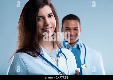 Female doctor is smiling with a male doctor out of focus behind her, both are wearing blue stethoscopes and white coats Stock Photo
