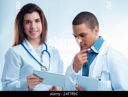 Two young doctors are using digital tablets while wearing white coats and stethoscopes. They are concentrating on the information on the screen Stock Photo