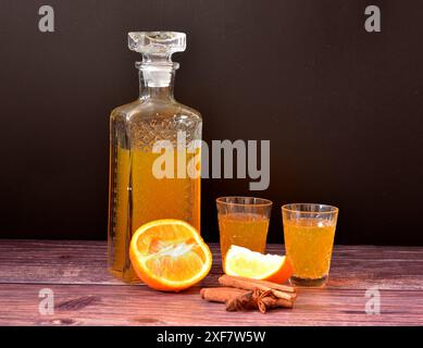 Homemade orange liqueur in a crystal decanter and two glasses on a dark wooden table, next to cinnamon sticks and star anise. Close-up. Stock Photo