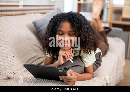 A happy, pretty young Black woman lying down on a couch in the living room and using her digital tablet, watching a movie, reading something online, o Stock Photo