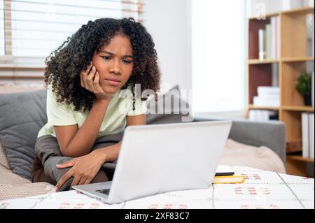 A serious young Black woman is sitting on the couch at home, reading an email or an article on a website, showing an uncertain, confused expression wh Stock Photo