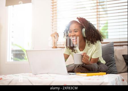 A cheerful young Black woman wearing headphones is sitting on a couch at home, showing her fist, excited and surprised by good news on her laptop. got Stock Photo