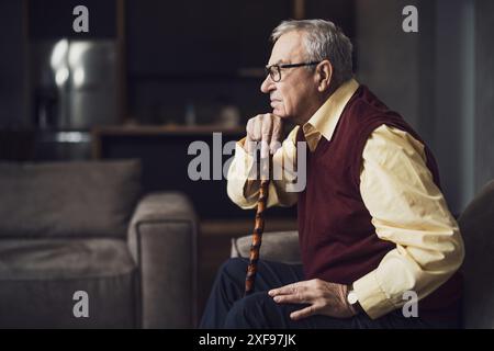 Pensive and worried senior man is sitting on bed in his home. Portrait of senior man with walking cane. Stock Photo