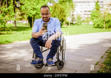 Portrait of happy man in wheelchair. He is enjoying sunny day in city park and messaging on smartphone. Stock Photo