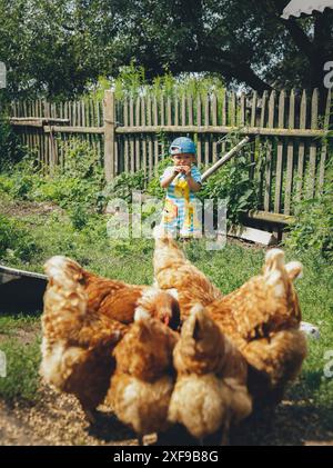 A child in bright summer clothes stands in a garden near a wooden fence, with a group of chickens pecking at the ground nearby.Belarus, Minsk Stock Photo