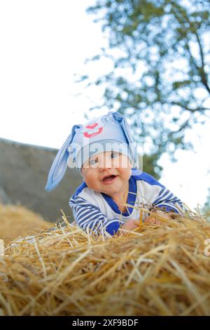 Smiling baby wearing a blue hat with bunny ears lying on hay outdoors with trees in the background.Belarus, Minsk Stock Photo