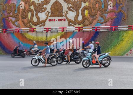 Locals riding motorbikes mopeds along busy road by mosaic wall in Hanoi, North Vietnam, Asia in June Stock Photo