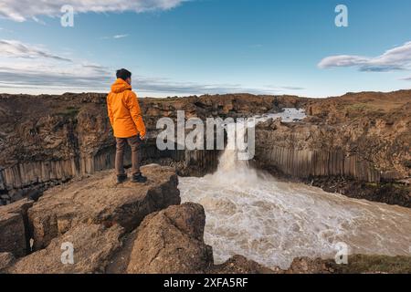 Traveler man in orange jacket standing and enjoying with powerful Aldeyjarfoss waterfall flowing and basalt column among the Icelandic Highland in sum Stock Photo