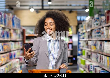 Woman smiling and using her smartphone while shopping in a grocery store aisle. She looks happy and engaged, pushing a shopping cart filled with items. Perfect for concepts lifestyle, and retail. Stock Photo