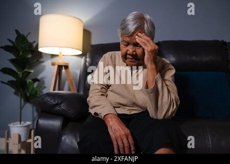 senior woman having a headache pain while sitting on a sofa in living room at night Stock Photo
