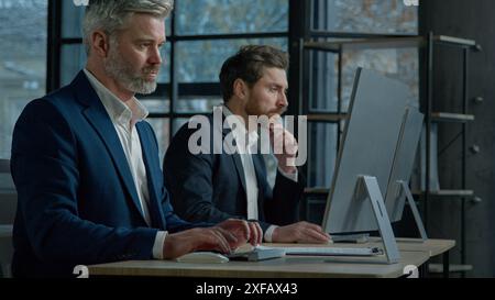 Focused professional managers working with computers on project sit in row at desk in office caucasian adult business men businessmen corporate Stock Photo