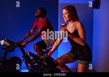 Biracial couple doing cardio in cardio neon zone of premium fitness club, woman smiling widely while she drinking BCAA, and man smiling while riding exercise bike Stock Photo