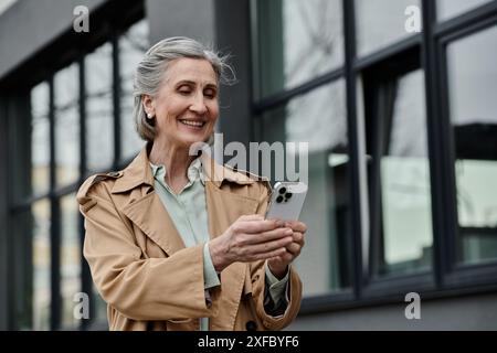 A stylish woman in a trench coat confidently holds a cell phone. Stock Photo