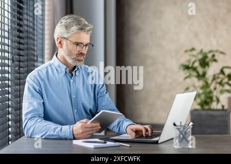 Mature office worker in a blue shirt working diligently on a laptop and tablet in a modern work environment. The scene portrays focus, concentration, and professionalism. Stock Photo