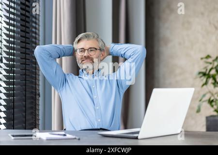 Mature male office worker taking a break with hands behind head, sitting at desk with laptop in a modern office. The man is wearing glasses and a blue shirt, looking content and relaxed. Stock Photo