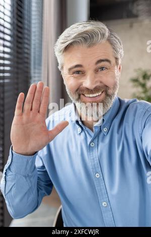 A cheerful mature office worker waves at the camera, greeting on a video call. Wearing a blue shirt, he presents a warm and friendly demeanor in a professional office setting Stock Photo