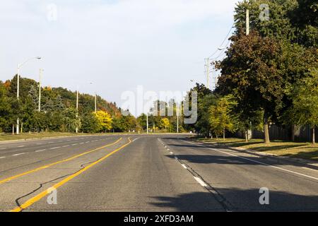 Sunlit suburban road - surrounded by green trees and power lines - blue sky - no vehicles or pedestrians. Taken in Toronto, Canada. Stock Photo