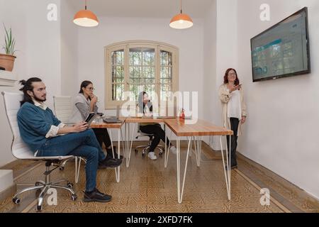 A professional redhead woman makes a statistical presentation to her diverse team using a large flat screen in a bright office environment. Stock Photo