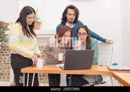 A group of young creative professionals working together at a desk in a bright office, looking at the laptop screen attentively. Stock Photo